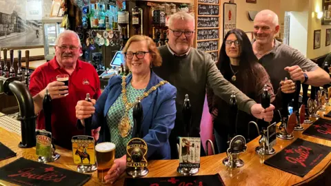 BBC Two women and three men standing behind a bar at a pub. Some are holding the beer pumps while others are holding drinks. One of the women is wearing a mayoral chain.