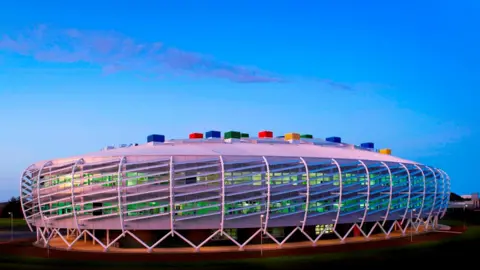 LDRS Colourful, oval-shaped, futuristic-looking Monkseaton High School which has multi-coloured blocks on its roof. The building is wrapped by white steelwork which sits on A-shaped white steel rods. Lights visible through windows make the rooms appear green coloured.