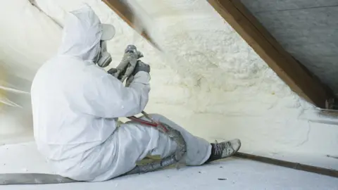 Getty Images Tradesman sits on the floor of a loft space, using tools to apply spray foam insulation to the underside of the roof while wearing a white set of overalls and a respiratory mask