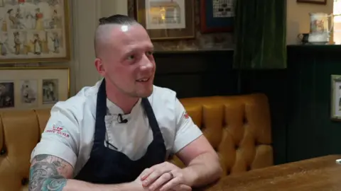 Chef Luke Emmess sitting on a table inside a pub during an interview. He is smiling and looking away from the camera.