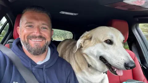 A man with short grey hair and a grey beard wears a navy blue hoodie in a car with red seats while sat next to a dog. 