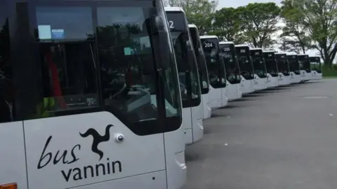 A line of modern silver buses. with large windows, text on the side reads bus vannin, they are in a large carpark.