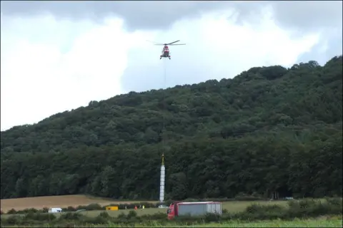 A helicopter hovers above the ground with a wire attached to a long white structure of about 15 metres. A number of people and vehicles are stationary in the field from where it is being lifted. 