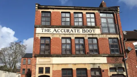MOLA A three storey building built in red brick with three rows of brown framed windows. Between the first and second floor windows is painted The Accurate Boot. 