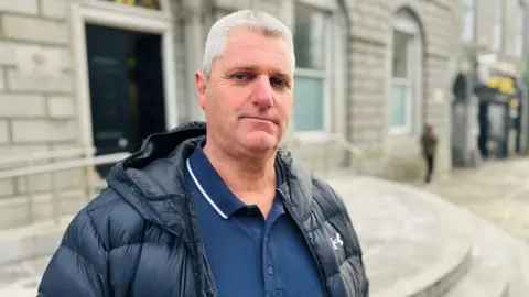 Ronnie Johnson, brother of victim - smiling at the camera outside court building. He has grey hair and is wearing a black quilted jacket and a blue T-shirt with a collar.