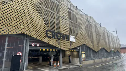 The image shows the exterior of a multi-story car park named Crown Car Park. The building features a distinctive architectural design with a facade made of gold-coloured lattice panels arranged in a geometric pattern, creating a textured and modern appearance. The panels are mounted on a metal framework that curves slightly, giving the structure a dynamic look.
At the entrance, there is a large black sign with bold letters spelling CROWN, accompanied by a smaller white sign that reads CAR PARK. The entrance is wide enough for two lanes, with yellow and black clearance bars overhead indicating height restrictions. Several traffic signs are visible, including speed limits and warnings, and two orange-and-white traffic cones are placed near the entry point. Inside the entrance, a dark-coloured vehicle is partially visible, along with ticket machines and barriers for controlled access.