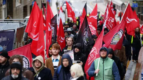 A group of people walk down the street as the protest. Many are holding red Unite or black flags.