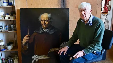 Wentworth Woodhouse Preservation Trust A man with short grey hair, wearing a green jumper and jeans is sitting in an artist's studio. To the left of him is an oil painting which shows a man in a brown gown with a white sash tied around the waist. The man in the painting is balding on the top of his head with grey hair to the sides. 