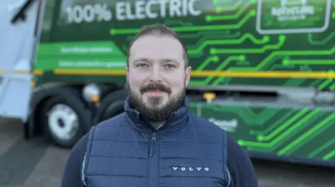 Mike Gill from Volvo wearing a navy blue Volvo gillet, smiling in front of the electric bin truck.