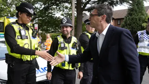 Reuters Andy Burnham, a man with dark hair, black glasses, and wearing a dark coloured blazer and white shirt, shakes hands with a police officer in uniform while three others look on. A police car can be seen in the background, as well as houses and trees.