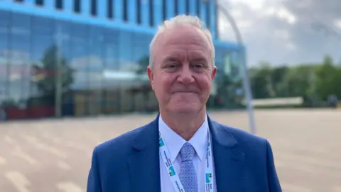 Laura Coffey/BBC A man in a blue blazer, light blue shirt and blue tie looks at the camera. A glass office building is behind him.