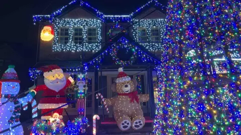 Paul Molyneux A house is covered in bright Christmas lights, with glowing decorations including Santa, a snowman, and a large teddy bear holding a candy cane. A tall tree wrapped in multicolored lights stands in the foreground. The scene is vibrant and festive against the night sky.