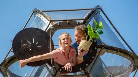 Woman and man inside a glass capsule looking out of a porthole type window. The woman has blonde hair and is wearing a short-sleeve pink shirt; the man is wearing a blue top and has short blonde hair and is holding a plant with a wicker pot.
