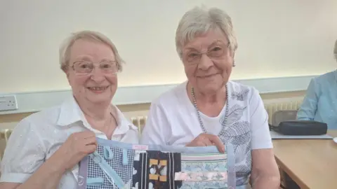 LDRS Two women with grey hair and glasses, both wearing white blouses and holding a piece of embroidery in their hands
