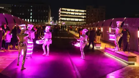 North News Pictures Street performers are lit up in pink light while playing the drums to people walking past.