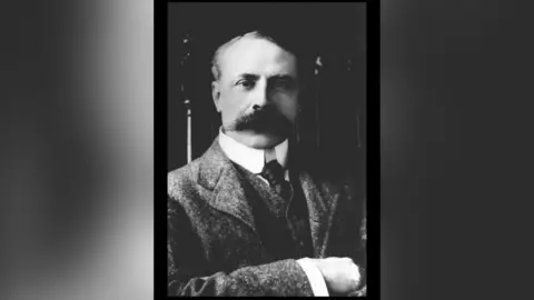BBC A black and white photo of Sir Edward Elgar wearing a grey suit. He has swept back short hair and a long, thick moustache.