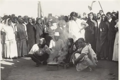 Alkazi Collection of Photography Three women and two men are seen crouched on the shores of Mumbai's Chowpatty Beach, ready to make contraband salt. A crowd, mostly comprising women, stands behind them and watches on. 