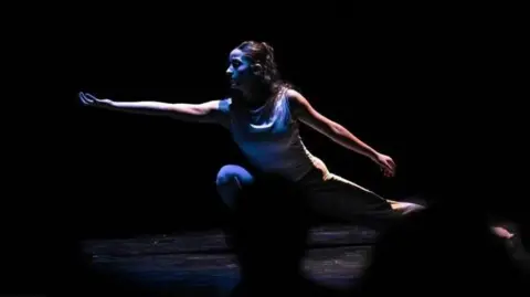 A contemporary dancer onstage, reaching with her hand turned towards the ceiling; one of her legs is in a deep squat (stock image)