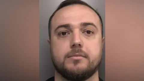 Merseyside Police A mugshot of Hani who is looking directly toward the camera. He has short, dark hair that is closely trimmed at the sides and slightly longer on top. His forehead is clearly visible, and the eyebrows are dark and moderately thick. Facial hair is visible around the jawline, chin, and upper lip, forming a short beard and moustache. The beard appears darker and slightly fuller around the chin area. The lips are closed, and the facial expression is neutral.