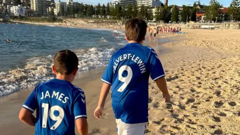 Family photo The back of a young Mackenzie and his younger brother wearing blue Everton jerseys with the names of players Dominic Calvert-Lewin and James Rodriguez as they walk on a sandy beach in Australia.