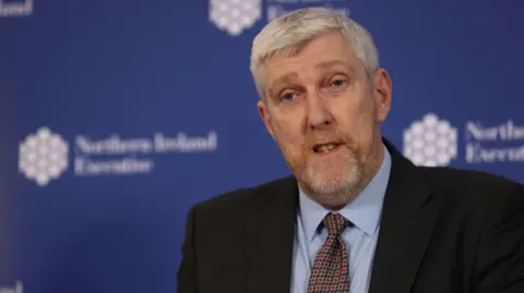 Finance Minister John O'Dowd, with grey hair and stubble. He is wearing a light blue shirt, a navy tie with red spots and a black suit jacket. There is a dark blue wall in the background. 