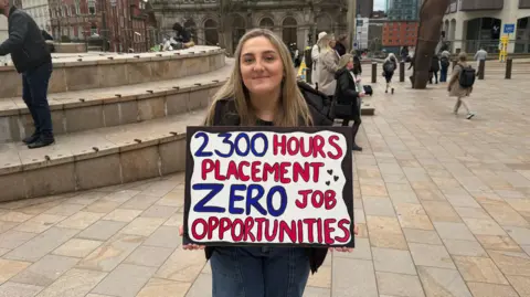 A woman with long blonde hair dressed in a black coat with blue jeans stands facing the camera holding a placard which reads: "2,300 hours placement, zero job opportunities". She is in a square next to some steps. There are people walking in the background.