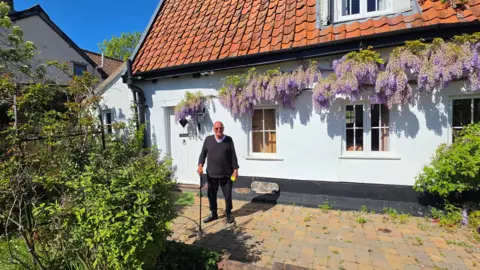 Supplied An older man with a grey jumper and black trousers on holding a walking stick. He is standing in front of a picturesque white cottage which has flowers hanging down from it. 