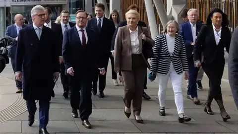 PA Media A group of people in business wear walk along a pavement 