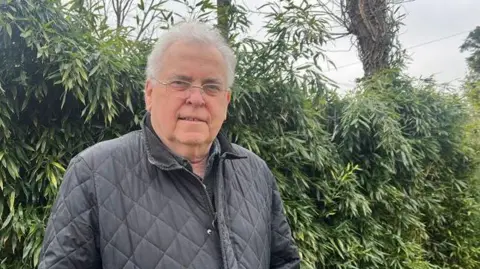 Peter Humphrey is standing in front a fence and shrubs in his garden with trees in the background. He has grey hair and glasses and is wearing a black jacket.