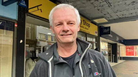 BBC Michael Hilderley smiles at the camera as he stands outside a shop front on a pedestrianised street. He is wearing a grey fleece, grey jumper and black T-shirt. He has grey hair. 