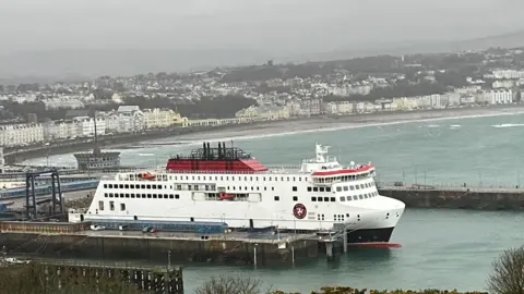 The Manxman ferry, which is white, red and black, moored in Douglas Harbour on an overcast day with Douglas Promenade in the background.