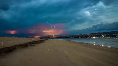 Ian McMichael Shows a derelict beach with water to the right and dark skies lit up by lightning 