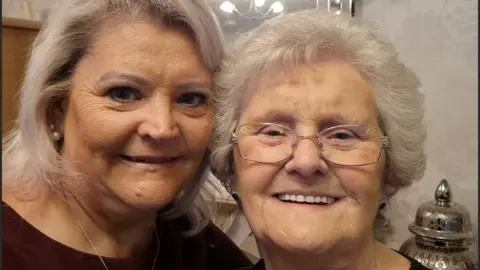 Handout Julie Harrison, who is on the left and wearing a purple shirt and a pearl style earring, is standing next to her mother, Joan, who is wearing thin silver rimmed glasses and is smiling at the camera. 