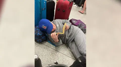 Jane Davis A pupil, in grey tracksuit and blue cap, is laying on the airport and reading a book. He is surrounded by suitcases and backpacks. 