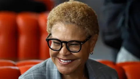 This is a head and shoulders pic of Karen Bass who is smiling broadly and wearing a grey, checked suit. The background, slightly out of focus, appears to be red seating