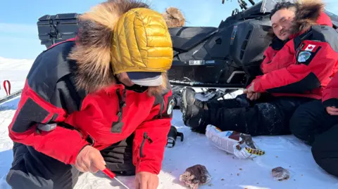 Nadine Yousif/BBC A man in a yellow hood and red parka cuts meat while another sits next to a snowmobile and watches