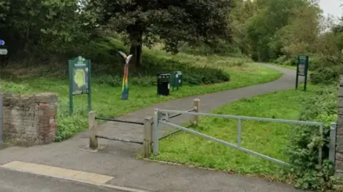 Google The entrance to a park with a low gate, two information boards and a path leading away between trees.