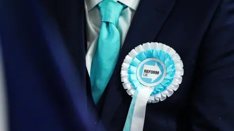 A Reform UK candidate standing with a blue and white rosette and blue tie.