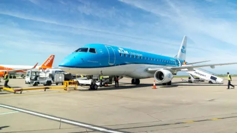 A blue KLM branded plane is seen on the ground at an airport runway, with a white and orange EasyJet plane in the background and airport workers in high vis jackets walking by.