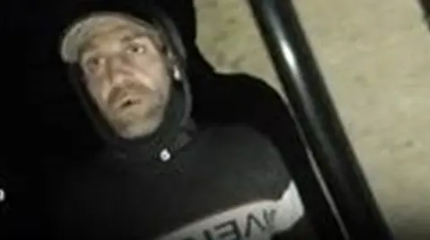 A blurry image of a man in a black hoodie. He seems to have a cap and light stubble.
