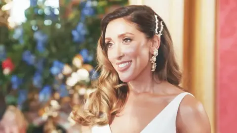 Married at First Sight UK, CPL Productions Shona stands in an elegant setting, wearing a white sleeveless wedding dress and ornate dangling earrings. She is captured side-on and smiling at her groom, who is off-camera. Her wavy light‑brown hair is styled with decorative pins on one side. Behind her, there is a blur of colourful flowers — blues, whites, golds and reds.