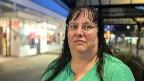 BBC/Elizabeth Baines Ms Dominic wears glasses and a green blouse. She has long black hair and a fringe. The post office can be made out in the background.