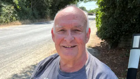 Mousumi Bakshi/BBC A man wearing a grey T-shirt and smiling. There is an empty road behind him.