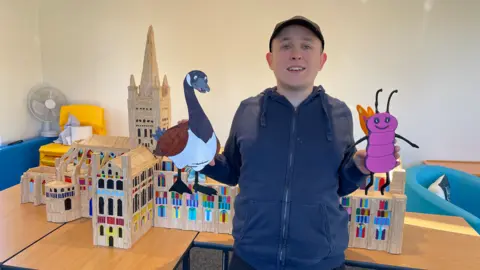 Edd Smith/BBC A man in a navy zip-up top and baseball cap standing in front of a model of Norwich Cathedral made out of matchsticks 