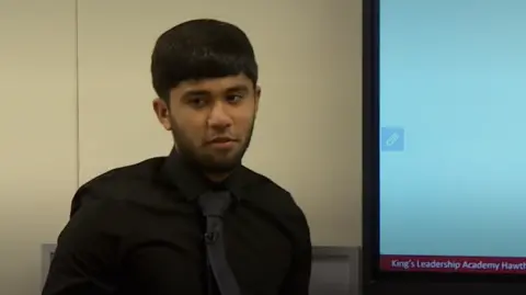 A young Asian man with black hair and a light black beard teaches a class. He is standing next to a digital whiteboard with mathematical equations written on it, and is wearing a black shirt and black tie.