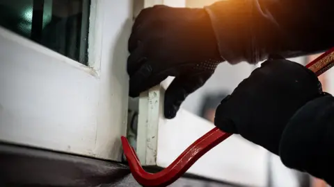 A person wearing dark gloves is prising open a window using a red crowbar or jemmy. The image shows only the hands, the tool and the white window frame