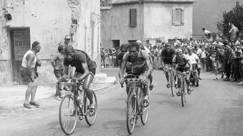 A black and white image of the Tour de France race in 1950. Cyclists can be seen making their way through a French town as spectators cheer them on.