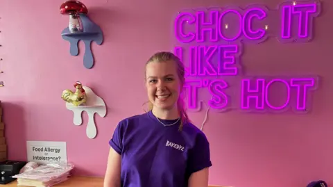 A woman with blonde hair smiles at the camera. She wears a t-shirt bearing the name of her business "Baked PZ". She is standing in her bakery shop. Behind her is a purple neon sign reading "choc it like it's hot".