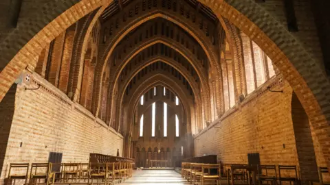 Getty Images A picture looking into Quarr Abbey. Chairs are lined up on either of the long hall. The ceiling arches overhead. Sun is bursting into the abbey through the windows to the right. 