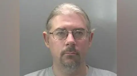 Robert Norman is looking at the camera in a police custody shot. He has fair hair that appears to be tied back, and a beard and moustache. He is wearing spectacles and a grey top.